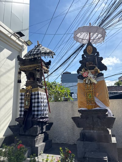 This image captures two Balinese temple shrines, likely located in Kuta Selatan, Indonesia, under a bright blue sky with numerous electrical wires overhead. The shrine on the left is adorned with a black and white checkered fabric, a traditional Balinese textile known as a "poleng." It is topped with a tiered, dark stone structure and a checkered umbrella. The shrine on the right is draped in yellow fabric, also a common sight at Balinese religious sites, and is similarly topped with a dark stone structure and a white umbrella. Both shrines feature decorative elements and offerings. The surrounding environment includes a white wall on the left and a textured, light-colored wall behind the shrines, with glimpses of green foliage and modern buildings in the background. The overall scene suggests a functional religious area within an urban or semi-urban setting.