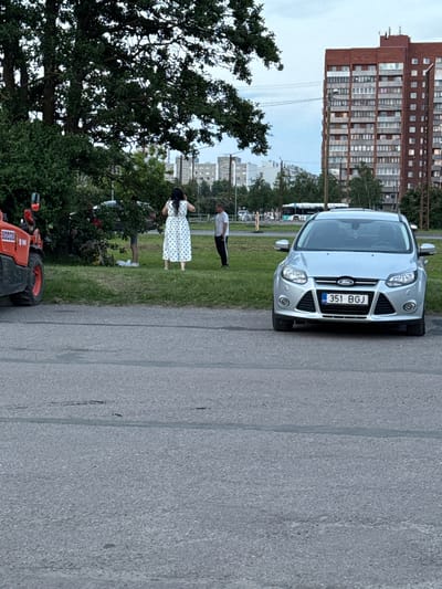 A silver Ford Focus, license plate 351 BGJ, is parked on asphalt in front of a grassy area. To the left, the orange front loader of a piece of construction equipment is visible. In the grassy area, a woman in a white and blue polka dot dress stands with her back to the camera, facing away from the car. Two other individuals are standing near some bushes to her left. Further back, a bus and several buildings, including a tall brick apartment building, are visible. The scene appears to be in an urban park or recreational area in Tallinn, Estonia. The sky is overcast.