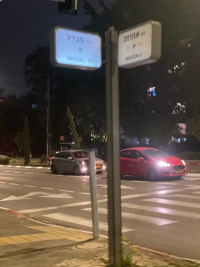A street corner at night with two lit street signs reading "Migdal St." and "Ahuza St." A red car is parked on the street to the upper right. A gray car with headlights on is parked at the intersection. The street has crosswalk markings and leads into the darkness. A building with a manhole cover is on the right.