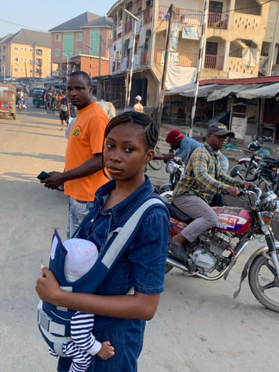 A daylight street scene depicts a bustling urban environment in Aba, Nigeria. In the foreground, a young Black female, with braided hair, wears a denim jumpsuit and faces the camera directly, holding an infant in a navy blue baby carrier strapped to her chest. The infant wears a light cap and striped sleepsuit. Immediately behind her, a Black male in an orange t-shirt and light-colored jeans stands, holding a dark smartphone in his left hand, also looking towards the camera. To the right, a Black male wearing a plaid shirt and dark baseball cap sits astride a red and silver Honda motorcycle, looking forward.
The background features a paved street with other pedestrians, motorcycles, and a green tricycle (Keke Napep) visible further down. On the right, a multi-story building with light-colored walls and balconies is visible, with multiple banners or fabric pieces hanging from it, and what appear to be ground-level market stalls. Further left in the background, additional multi-story buildings with light and orange facades are present. Utility poles and overhead wiring are visible throughout the scene. Shadows cast on the ground suggest a specific time of day with natural sunlight.