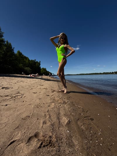 The image depicts a sunny day at a sandy beach in Rusanovo, Russia, adjacent to a calm body of water, likely a river or large lake. A prominent young woman stands barefoot in the foreground on the damp sand at the water's edge, facing the viewer. She is wearing a bright neon green one-piece swimsuit and sunglasses, with long, light-colored hair, part of which is styled into dreadlocks. Her left arm is raised with her hand behind her head, and her right hand is placed on her hip, indicating a posed stance. Her body casts a long shadow on the sand behind her.
In the mid-ground, further down the sandy beach to the left, several other individuals are present. One person is distinctly visible lying supine on a light-colored towel or mat on the sand, wearing a bikini. Other figures are scattered along the treeline and closer to the water. The background on the left is dominated by a dense line of dark green deciduous trees. To the right, the wide expanse of water extends to a distant forested shoreline under a clear, deep blue sky with minimal white clouds. The sandy beach shows various textures, footprints, and scattered small natural debris like sticks.
