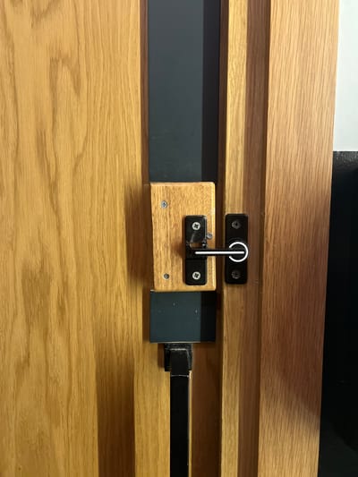 A close-up shot depicts a wooden cabinet or door featuring a sliding bolt lock mechanism. The wood is light-colored with a visible grain. The lock consists of a black metal bracket fixed to the wood and a wooden block secured by screws. A white and black handle is attached to the wooden block, allowing it to slide along a black metal rail. The rail extends horizontally across the frame. The environment consists of the immediate surface of the wooden structure and some darker material above it, possibly the top of the cabinet. The location is not discernible from this image.