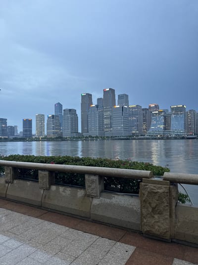 The image depicts a city skyline at dusk, with a serene body of water reflecting the illuminated buildings. The perspective is from a low vantage point, looking across the water at a dense collection of modern skyscrapers. The buildings are of varying heights and architectural styles, with many featuring glass facades that catch the fading light. Some buildings display corporate logos or names, though they are not clearly legible.
The time of day is clearly evening, as indicated by the deep blue of the sky transitioning into darker hues, and the artificial lights of the city beginning to dominate. The water's surface is calm, with gentle ripples creating distorted reflections of the cityscape.
In the foreground, a stone embankment with decorative carvings forms a barrier, separating the viewer from the water. There are hints of greenery and small pink flowers growing along the embankment, adding a touch of nature to the urban scene. The ground in the immediate foreground is paved with large, light-colored tiles.
The overall mood of the image is peaceful and contemplative, with the soft lighting and still water creating a sense of tranquility. The scene suggests an urban waterfront area, likely a promenade or park, designed for public enjoyment of the city's architecture and natural surroundings. The location is contextually identified as Haizhu District, China. No people are visible in the frame.