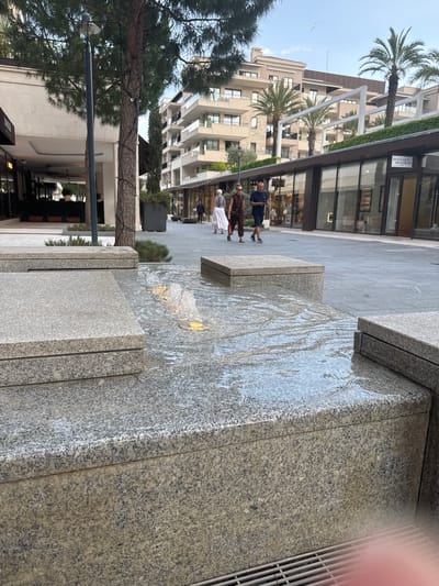 The image depicts an outdoor scene with a modern architectural setting. In the foreground, a tiered water feature made of granite is visible, with water cascading over its surfaces. The stone has a speckled, grey and black pattern. A metal grate is also present on the right side of the foreground.
In the midground, several people are walking on a paved walkway. One person, a man in a dark blue shirt and shorts, is facing left. Two other individuals are further down the path. The walkway is bordered by buildings with large glass windows and modern facades, suggesting a commercial or residential area. Palm trees are interspersed with the buildings, indicating a warm climate.
The background features multi-story buildings with balconies and light-colored exteriors. The sky is visible above, appearing overcast or early evening. The scene appears to be in a developed urban area, possibly a plaza or promenade.
There is text visible on a building in the upper left portion of the image, which appears to read "BIOLOGIQUE RECHERCHE".
The overall impression is one of a clean, contemporary urban environment, possibly in Tivat, Montenegro, with people enjoying the public space. The time of day is likely late afternoon or early evening.