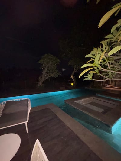 The image depicts an illuminated outdoor swimming pool area at night in Batuan, Indonesia. A rectangular swimming pool, featuring an integrated square-shaped jacuzzi or plunge pool, is illuminated with vivid blue light. Dark wooden decking borders a section of the pool, upon which a white-framed outdoor lounge chair with dark cushions is positioned in the foreground. A partial view of a white, irregularly-shaped outdoor table is adjacent to the lounge chair. Multiple trees with green foliage are visible against the dark sky in the background, with sparse warm-toned artificial lighting present among them. A large plant with prominent light green leaves partially frames the upper right portion of the scene. No individuals are visible.