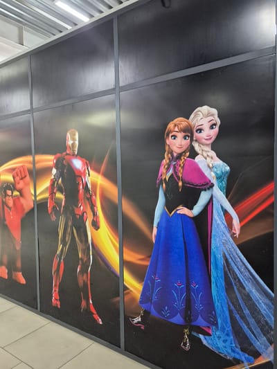 This media file showcases a ceiling mural featuring characters from popular animated and superhero franchises. The top section of the mural depicts Elsa and Anna from Disney's "Frozen." Elsa, with her signature blonde braid and ice-blue gown, is positioned behind Anna, who has auburn hair and is wearing a royal blue dress with a pink and black bodice. Their expressions are friendly and engaging, suggesting a cheerful and magical scene.
Below the "Frozen" characters, the mural transitions to feature Iron Man from the Marvel Cinematic Universe. The armored superhero is depicted in a dynamic pose, seemingly in mid-flight with trails of orange and yellow energy emanating behind him, symbolizing speed and power. Further down, the red fist of what appears to be another superhero is visible, along with a portion of a character's face, possibly from a different Marvel film or franchise.
The setting appears to be an indoor public space, as evidenced by the ceiling panels and the glimpse of a tiled floor in the lower right corner. The lighting suggests daytime, but it's difficult to ascertain precise time or weather conditions due to the indoor location. The overall impression is one of a vibrant and exciting environment, likely a place geared towards children or fans of these fictional characters, such as a shopping mall, entertainment venue, or themed area within Kokshetau, Kazakhstan. There is no visible text in the media file.
