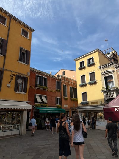 This is a photograph of a street scene in Venice, Italy. The image depicts several buildings with distinct Venetian architecture, characterized by their colorful facades (yellow and reddish-orange) and shuttered windows. In the foreground, a cobblestone pavement is visible, suggesting a pedestrian area.
Several people are present in the scene, walking along the street or in front of shops. On the left, a person in a blue shirt and pink shorts walks away from the camera. In the center, two women are walking towards the camera, one with long dark hair wearing a white off-the-shoulder top and black shorts, and the other wearing a dark top and dark skirt. A man in a black t-shirt and jeans is walking away from the camera on the right. Other individuals are visible further in the background, in front of shops and restaurants.
Various shops and businesses are identifiable. A shop with a white awning and displays of merchandise is on the left. A restaurant or trattoria with green awnings and outdoor seating is visible in the middle, with a sign reading "trattoria Vittoria". Another establishment with a dark red awning is on the right, with text partially visible. The overall atmosphere suggests a lively commercial area in Venice. The sky above is blue with scattered clouds.