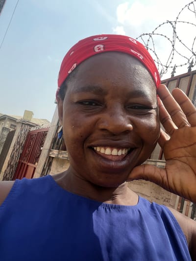 The image is an outdoor, close-up selfie of an adult female, depicting a scene in Iranla, Nigeria. She has dark skin and is smiling broadly, revealing her teeth. Her head is covered by a red fabric headwrap adorned with white patterns. She is wearing a royal blue sleeveless top, and a blue drop earring with a metallic detail is visible on her left ear. Her right hand is partially shown, positioned near her jawline with fingers slightly spread. The background features a clear blue sky with sparse white clouds. Visible urban elements include a light-colored building, a brown metal gate, and a textured wall on the left side. On the right, a darker fence or gate is topped with multiple coils of razor wire.