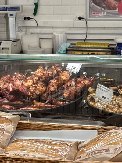 The image shows a close-up view of a display case filled with grilled meat skewers and bread. The meat skewers, some appearing to be chicken and others pork, are seasoned and cooked to a golden brown. They are arranged vertically on metal rods, suggesting they are part of a rotisserie or a similar cooking setup. To the right of the meat, several loaves of bread are visible, wrapped in clear plastic packaging with brown paper accents. These loaves are housed in a wicker basket, adding a rustic touch to the display.

In the background, a section of the store's wall is visible, tiled in white. A black electrical cord is connected to a white wall socket, indicating the presence of power for the display. There are also stacks of white plastic containers and a green-handled knife on a shelf. A price tag is visible on one of the meat skewers, with the text "149 90" printed on it, likely indicating the price in rubles. Another tag above the bread displays "99 90". The overall setting appears to be a food retail environment, possibly a supermarket or a deli counter, given the presence of food items and display fixtures. The lighting is bright, suggesting it is daytime or the store is well-lit. There are no people visible in the image, focusing solely on the products. The brand "SPAR" is visible on some of the packaging, identifying the store or brand. The image seems to be taken from a slightly elevated angle, looking down into the display.