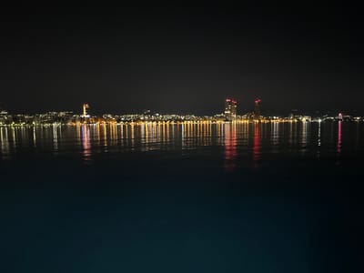 The image shows a nighttime view of the Limassol skyline in Cyprus, as seen from across the water. The city lights are reflected in the dark, calm water, creating a shimmering effect. Several buildings, including tall skyscrapers, are visible along the coastline, illuminated by a mix of white, yellow, and red lights. The sky above is dark and clear, with no visible stars or moon. The overall scene depicts a peaceful and picturesque urban landscape at night.