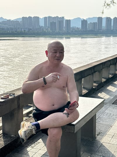 An older man, bald and shirtless, sits relaxed on concrete steps beside a body of water in Jiangjin, China. He wears dark shorts, light-colored sneakers with blue socks, and has a dark beaded bracelet on his left wrist and a red bracelet on his right. He holds a lit cigarette in his right hand, bringing it to his mouth. Keys and a red object, possibly a phone or lighter, are visible on the steps near his right leg.

The man's posture suggests he is leaning back, enjoying the moment. Behind him, a wide river stretches out, its surface shimmering brightly under the sunlight. Across the river, a dense skyline of numerous tall buildings rises, partially obscured by a light haze. A few green leaves or branches are visible on the far left, framing the river and city view. The steps he sits on appear to be part of a promenade or riverside embankment, constructed from a light-colored, patterned stone or concrete. The strong, direct sunlight casts clear shadows, indicating a bright and sunny day. No discernible text is visible in the image.