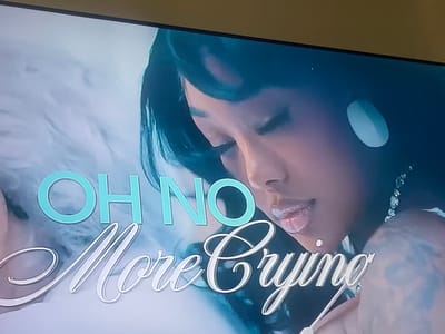The image displays a woman with dark hair, fair skin, and a diamond necklace. Her eyes are closed, and she has an earring in her ear. The text "OH NO More Crying" is overlaid on the image. "OH NO" is in sans-serif font and a blue-green color, while "More Crying" is in a cursive font and white with a light brown shadow. The background appears to be a light, marbled pattern.