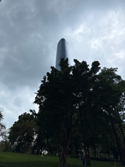 This is a low-angle, medium shot of a curved, glass-facaded skyscraper partially obscured by the dense foliage of trees in the foreground. The sky is overcast with dramatic, grey clouds. The skyscraper, identified as being in Tianhe District, China, appears tall and modern, with a sleek, reflective surface. The trees are dark green and leafy, creating a stark contrast with the building. The lower part of the image shows a green lawn, and a person in blue overalls is visible in the distance, seemingly engaged in groundskeeping. The overall atmosphere is moody and somewhat dramatic due to the lighting and weather conditions.