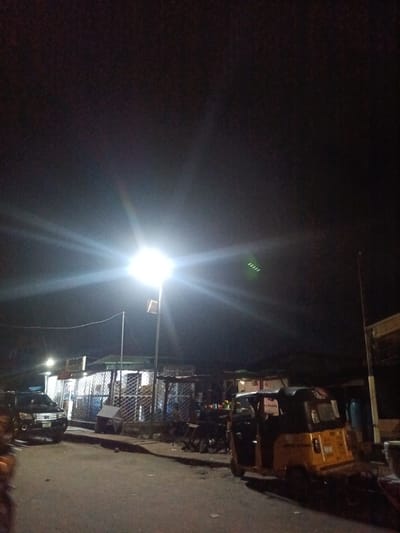 The image depicts a nighttime street scene in Ado, Nigeria, illuminated by a bright street lamp. A black SUV is parked on the left side of the road. Further down the street, a yellow auto-rickshaw (Keke Napep) is visible. Several small market stalls with metal grates are set up along the right side of the road. The sky is dark, indicating it is late at night. There are no discernible people actively engaged in actions, though figures may be present within or near the stalls and the auto-rickshaw, obscured by the darkness and distance. The environment appears to be an urban or semi-urban street with basic commercial structures.