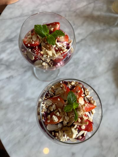 The image displays an overhead view of two identical dessert parfaits served in clear stemmed glasses, positioned on a polished light gray and white marble table. Each parfait features layered components, including a dark reddish-purple fruit or berry mixture at the base and a creamy white layer. The desserts are generously topped with slices of fresh red strawberries, a mixture of chopped nuts predominantly consisting of slivered almonds, and some coarser fragments consistent with walnuts. A single sprig of fresh green mint garnishes the center of the topping in both glasses. The scene is static and appears to be set indoors, likely within a cafe or restaurant environment in Uluwatu, Indonesia. No people are visible.