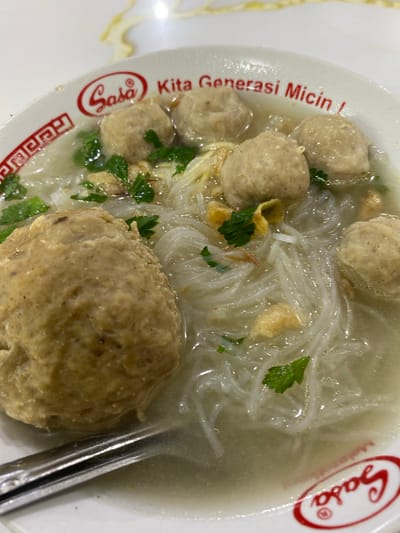 This is a close-up, top-down shot of a bowl of Indonesian bakso, a meatball soup. The bowl is white with red accents and features text that appears to be in Indonesian. The soup contains several large, light brown meatballs, thin white noodles, and some green herbs, likely cilantro or parsley, as a garnish. A metal spoon is partially submerged in the broth on the right side of the bowl. The lighting suggests an indoor setting, possibly a restaurant. The text on the bowl is partially visible and includes "Gasa" within an oval logo, and also "Generasi Micin". The overall presentation is appetizing, focusing on the texture and ingredients of the dish. The setting appears to be a casual dining environment, fitting for a popular street food like bakso. No people are visible in the image.