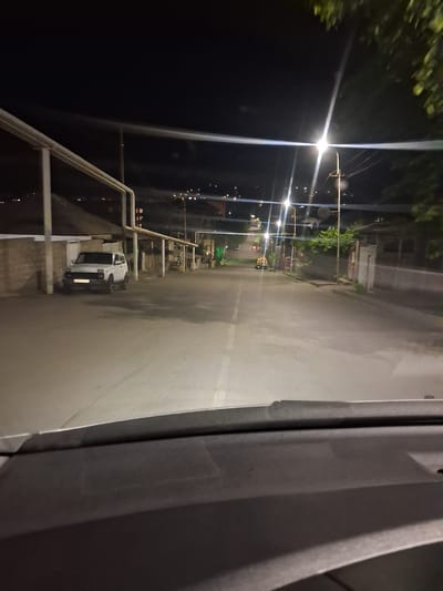The image depicts a street scene at night in Yeghegnadzor, Armenia. A white SUV is parked on the left side of the road, facing towards the viewer. The street is paved and illuminated by streetlights. Buildings and trees are visible on either side of the road, and in the background, distant lights suggest a town or city. The perspective suggests the photo was taken from inside a vehicle.