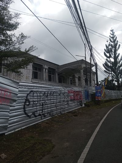 An unfinished, large building with classical architectural details sits behind a corrugated metal fence covered in graffiti. The overcast sky and bare trees suggest a bleak or transitional season. A sign, seemingly an advertisement, is placed along the fence near the edge of a narrow road, partially obscured by vegetation. Power lines cross the sky above the scene.