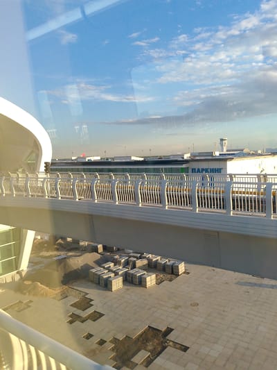 This image captures an outdoor scene, likely at an airport or a similar large transportation hub, as suggested by the presence of a control tower and the word "ПАРКИНГ" (PARKING) visible on a building. The scene appears to be under construction, with piles of sand and neatly stacked construction materials on the ground. Paving stones are laid out on the ground, with some sections missing, indicating ongoing work.

The foreground features a modern pedestrian bridge or walkway with white railings, casting a shadow on the ground below. The sky is blue with scattered white clouds, suggesting fair weather and potentially late afternoon or early morning, given the angle of the sunlight. Reflections are visible on the glass surface from which the photo was taken, obscuring parts of the background and sky. The architecture appears contemporary, with clean lines and large glass elements.

There are no people visible in the frame. The overall impression is one of a functional, modern infrastructure project in progress. The location cue of "Astana, Kazakhstan" provides specific geographical context. The prominent text "ПАРКИНГ" on the building directly identifies its purpose.