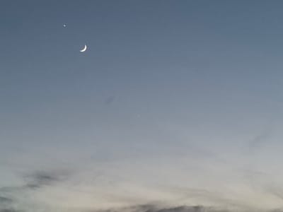 The image depicts a twilight sky with a crescent moon and two visible stars. The sky transitions from a lighter hue at the bottom to a deeper blue at the top, suggesting the transition from sunset to night. Wispy clouds are present towards the bottom of the frame, tinged with the fading light. In the upper left quadrant, a bright star is positioned above and slightly to the left of the crescent moon. Another fainter star is visible further down and to the right, closer to the center of the image. The scene evokes a sense of calm and the vastness of the night sky. There is no visible text in the image. The location context is Pchelnik, Bulgaria.