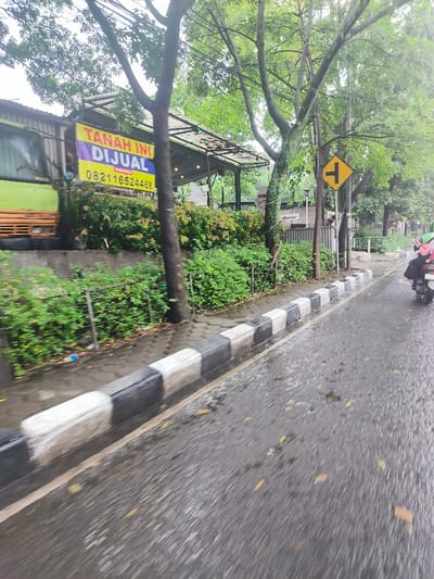 The image shows a wet road in Bandung, Indonesia, with a curb and a sidewalk on the left. A large tree is in the foreground, blocking a view of the road ahead. A yellow sign with a black "T" indicating a T-junction is attached to a post on the right side of the road. A person is driving a motorcycle towards the camera. A sign in Indonesian language advertises land for sale, with a phone number. The road is wet from recent rain, and there is a small amount of vegetation on the side of the road. A large green vehicle is partly visible on the left side of the image. The curb is made of black and white concrete blocks. The image is blurry, likely taken by a person moving or riding in a vehicle.
