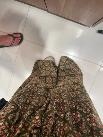 The image shows a first-person perspective of two legs wearing a green patterned skirt and one foot wearing a dark grey flip-flop. The person is standing on a white tiled floor. In the background, a wooden surface is visible, possibly a cabinet or countertop. The scene appears to be indoors, in a residential or commercial setting. There is no visible indication of the location being Mumbai, India.