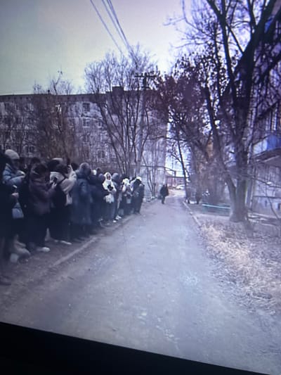 The image shows a long line of people standing alongside a path or road in an urban environment. Bare trees line both sides of the path, and a multi-story building is visible in the background. The scene appears to be taken during winter or early spring. The location is identified as Cheongju-si, South Korea.