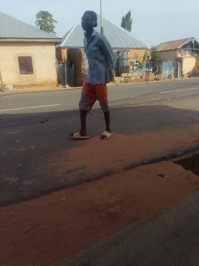 A full-shot medium outdoor photograph shows a man walking along a street in Tempure, Nigeria. The man is wearing headphones, a blue button-down shirt, and patterned orange shorts. He is wearing beige slides on his feet. In the background, there are several buildings with corrugated metal roofs, a dirt area, and a paved road. The sky is clear and blue.