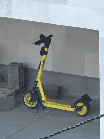 The image shows a yellow electric scooter parked outdoors in Tbilisi, Georgia. The scooter is leaning against a textured, grey concrete surface, which appears to be a wall or the side of a building. To the right of the scooter, a thick black and white cable runs diagonally down the frame, suggesting the presence of power lines or telecommunication infrastructure.

The scooter itself is bright yellow with black accents. Its handlebars are visible, with black grips and a black brake lever on the left side. The front wheel is dark, and the rear wheel, also dark, has a distinctive yellow rim with spoke-like cutouts. The body of the scooter is a prominent yellow bar, with some blurry yellow text visible on its side. A black fender is visible over the rear wheel.

In the background, a section of a light-colored, slightly textured surface is visible, possibly a wall or pavement. Below and to the left of the rear wheel, a grey, rectangular concrete block is partially visible, suggesting an urban environment. The lighting suggests it is daytime, though the weather cannot be determined. The overall scene depicts a typical urban setting in Tbilisi where electric scooters are commonly used for transportation.