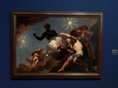 The painting, displayed in a dark wooden frame, depicts a celestial scene with mythological figures. In the center, a large, textured sphere is being manipulated by several beings. An older, muscular male figure with a beard and clad in a blue cloth is pushing against the sphere with his right arm, his body contorted in exertion. To the left, a cherubic figure with wings and a red cloak rides on a white swan, holding a torch that emits smoke. Behind the sphere, a dark-skinned male figure with a headdress made of red feathers is visible, also seemingly interacting with the sphere. Above and to the right, a winged figure with a red cloak is reaching towards the sphere, holding what appears to be a cluster of small, round objects. The background is a twilight sky with prominent starbursts and dark, swirling clouds, suggesting a cosmic or divine setting. The painting is hung on a deep blue wall. To the right of the painting, a small white label is affixed to the wall, providing information about the artwork, including the artist Abraham Hondius (1628-1691) and the title "De overwinning van de tijd op de skulptuur" (The Victory of Time over Sculpture). This artwork is located in the Kunstmuseum Basel, Switzerland.