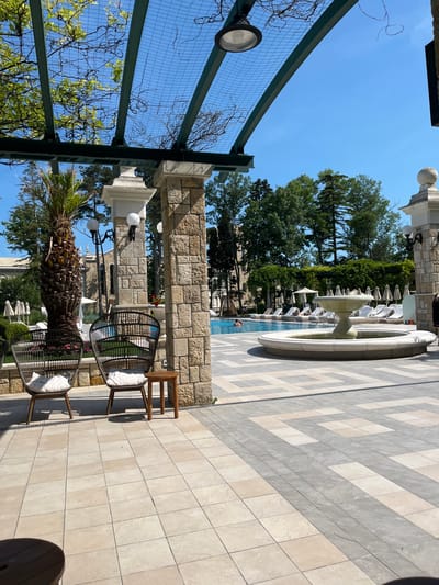 This image displays an outdoor resort or hotel pool area, identified as being in Varna, Bulgaria. The foreground features a tiled patio composed of light beige and grey square pavers in a geometric pattern. Two woven dark brown chairs with white seat cushions are positioned on the left, accompanied by a small, circular wooden side table between them. A large, rough-hewn stone pillar, constructed from irregular light brown blocks, stands prominently on the mid-right, supporting a dark green metal pergola overhead. To the left of this pillar, a slender white pillar is partially visible, adjacent to a thick-trunked palm tree with a robust, textured trunk.

The pergola above is made of dark green metal beams with a grid mesh, supporting sparse green foliage and dry vines. A single recessed ceiling light fixture is visible within the pergola structure. Several white globe light fixtures are mounted on the pillars and a black streetlamp-style pole further left.

In the midground, a blue swimming pool is visible, containing at least one individual on the left side. The pool is surrounded by numerous white lounge chairs, each paired with a matching white umbrella. To the right of the pool, a multi-tiered light-colored stone fountain is centrally located within the paved area. Dense green trees form the background, extending across the horizon beneath a clear, cloudless blue sky. A building with a reddish-orange tiled roof is partially observable through the trees in the far left background.
