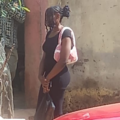 A woman with braids and a pink and white patterned backpack is outdoors in what appears to be Dadi Riba, Nigeria. She is wearing a black shirt and black leggings, and is holding what appears to be a cell phone. She is looking down at her phone, and there is a red car parked to the right. The background is a concrete wall, and there are some plants and debris on the ground.