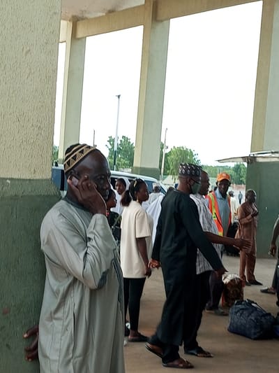 In Gombe, Nigeria, a group of people stand near a building with a textured green wall and exposed concrete pillars. A man in a grey traditional outfit and a patterned cap is on his phone, leaning against the green wall. To his right, a woman in a white outfit and black leggings stands looking towards the right. Further back, a man in a black traditional outfit and a face mask is interacting with another man wearing an orange cap and a reflective vest. Luggage, including a blue duffel bag and a patterned bundle, is scattered on the ground. The environment appears to be an outdoor waiting area, possibly a transport terminal, with glimpses of trees and a bright sky in the background.