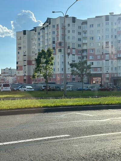 The image depicts a view from a moving vehicle, looking out over a residential area in Minsk, Belarus. The scene is dominated by a tall, multi-story apartment building with a facade featuring a grid of windows and balconies, punctuated by sections of light grey, white, and pinkish-red panels. A street lamp stands on the left side of the frame, extending into the sky.
In front of the apartment building, a strip of vibrant green grass runs alongside a paved road. Several cars are parked along the grass verge, including a white van, a blue car, and a dark-colored car. A young tree with lush green foliage is positioned in front of the building, casting a shadow on the grass. The sky is a clear, bright blue with a few fluffy white clouds visible on the left, suggesting fair weather. The sun appears to be high, indicating it is likely midday.
The perspective suggests the photo was taken from a slightly elevated position, possibly from another vehicle or a pedestrian overpass. The road in the foreground shows faint white lane markings. The overall impression is a typical urban residential scene in an Eastern European city on a sunny day. There is no visible text in the image.