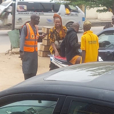 The image depicts a street scene in Dadi Riba, Nigeria. Several people are visible, including a man wearing an orange traffic vest, a woman with a headwrap and patterned yellow clothing, a man in a black shirt, and a person in a yellow shirt with "RTEAN KADUNA STATE" printed on the back. The people are gathered near several vehicles, including buses and cars. Boxes are visible near the woman, and papers are being handled by someone near the yellow-shirted person. The environment appears to be an outdoor street setting with trees in the background.