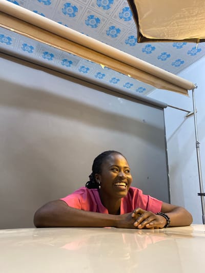 A medium shot shows a Black woman with braided hair wearing a pink t-shirt and cross earrings, resting her arms on a light-colored surface, her hands clasped in front of her. She is looking to her right and smiling broadly, with her teeth showing. In the background, a gray backdrop is visible, with a textured ceiling overhead featuring a blue and white patterned design. A light fixture or softbox is positioned above and behind her. The overall environment appears to be an indoor studio setup in Gura Top, Nigeria.