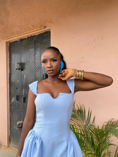 This media file features a young Black woman standing outdoors. She is wearing a light blue dress with ruffled details on the skirt and off-the-shoulder sleeves. She accessorizes with multiple gold bracelets on her left arm and large blue floral earrings. Her makeup is visible, with dark eyeliner and a glossy lip. Her left arm is bent, with her hand resting on her head.
The background consists of a textured, light pink wall with some graffiti, and a metal door or gate with a diamond plate pattern. To the right of the woman, palm fronds from a plant are visible. The lighting suggests it is daytime, likely outdoors. The overall mood appears to be a casual portrait or fashion shot.
Visible text on the wall includes "917838" and "027". Based on the provided location context, the setting is Gwong, Nigeria.