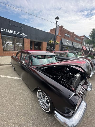 This is a photograph of a classic black car parked on a street in the City of Leduc, Canada. The car has a prominent chrome bumper and fender, and the hood is open, revealing the engine. The car's paint is a deep black with a subtle red glitter effect, and it features flame graphics on the hood. It has whitewall tires and chrome wheels.
The background shows a brick building with several storefronts. Visible text on the storefronts includes "SARADEE Boutique," "STUDIO," "gallery," "BLACKBIRD Knitting & Needlework," "RED DOOR ESTHETICS," and "SAGE SOL." A street lamp with hanging lights is also visible, as are flower baskets hanging from the lamp post. The sky is cloudy, suggesting it is daytime, possibly overcast. People are visible in the background, near the storefronts, indicating a public area, possibly a street fair or a shopping district. The overall scene suggests a car show or a downtown street on a day with some public activity.