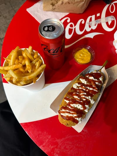 An overhead, close-up shot captures a Coca-Cola themed table featuring a can of Coca-Cola Zero Sugar, a cup of french fries, a small container of yellow mustard, and a corn dog drizzled with ketchup and mayonnaise. The table's surface is predominantly red with white accents, and a white paper napkin is visible in the upper background. The corn dog is served in a white paper tray, and the french fries are in a white paper cup. The scene appears to be set outdoors, possibly at a food stand or casual dining establishment. The lighting suggests it is daytime. The location is in Belgrade, Serbia.