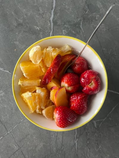 A white bowl with a yellow rim sits on a dark gray, mottled surface, possibly a countertop. Inside the bowl is a fruit salad consisting of sliced oranges, whole strawberries, and sliced peaches or nectarines. A thin, silver utensil, likely a fork or spoon, is resting in the fruit salad, with its handle extending out of the bowl. The lighting suggests an indoor setting. There is no indication of the city of Vologda, Russia, or any other specific location.