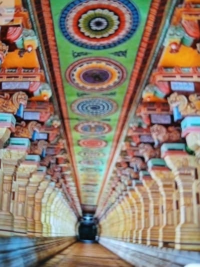 The image depicts the interior of a hall in a temple, likely located in Bengaluru, India. The walls are adorned with colorful circular patterns and sculptural figures. The ceilings appear to be carved and ornamented, creating a symmetrical and receding perspective. The hall seems long, leading to a dark area in the distance. There are no people visible. The environment suggests a place of worship with traditional Indian artistry.