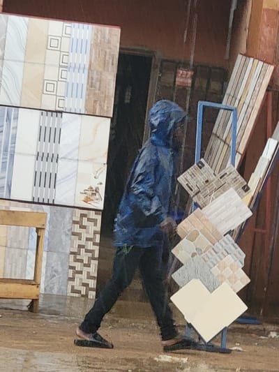 This vertically oriented image, captured in Maiburiji, Nigeria, depicts a person standing amidst a display of floor and wall tiles, likely within a building material store or an outdoor market stall.
The primary subject is a person, whose face is obscured by a dark blue hood, wearing a matching dark blue jacket or raincoat and dark pants. They are standing with their left leg slightly forward, facing towards the right side of the frame, seemingly observing the tile displays.
The scene is dominated by various ceramic or porcelain tiles. In the background, a series of smaller, patterned tiles are arranged on an angled blue metal display rack. These include tiles with intricate geometric designs, stone-like textures, and plain light colors. In the foreground, larger rectangular tiles are displayed vertically, showcasing a diverse range of patterns, including brick-like textures, horizontal stripes, abstract waves, and geometric squares.
The setting appears to be a practical, possibly open-air or semi-covered commercial space. The ground is a light, dusty brown. In the upper background, construction materials like wooden planks are visible, alongside what appears to be a blue tarp or bag. A dark doorway or opening is situated behind the person. A rough, brick-like wall section is also discernible in the background.
There is no specific activity evident beyond the person standing and observing. The time of day appears to be daytime, possibly overcast or shaded, as there are no harsh shadows. No distinct weather conditions can be inferred, though the hooded attire might suggest protection from elements.
A small, blurry red and white object with unreadable text is visible on a background wall section, but no other readable text is apparent. The overall impression is that of a bustling commercial environment focused on building supplies.