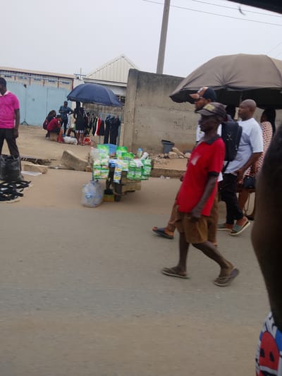 This candid daytime photograph captures a bustling street scene in Fegge, Nigeria, under an overcast sky. The image focuses on pedestrian activity and street vending.
In the immediate foreground on the left, a man in a pink polo shirt stands beside several black bags and a pile of black straps or sandals, possibly for sale. Further back, a wooden cart is laden with numerous packaged instant noodles or snacks and several bottles of water, indicating a mobile vendor.
To the left of the cart, under a dark blue umbrella, several people are gathered around a makeshift stall. Clothes are visibly hanging on a rack, suggesting a clothing vendor. One person is seated, wearing a red top. In the background on the left, light blue industrial gates and buildings with white roofs are visible.
On the right side of the frame, several individuals are walking from left to right. A man in a red polo shirt, brown shorts, and flip-flops is prominent in the mid-ground. He is followed by a man wearing a white short-sleeved shirt, dark trousers, and a black backpack. Another man in a black cap and glasses, and a woman in a patterned dress carrying a bag, are also part of this group. A large beige umbrella provides shade further down the street on the right.
The scene depicts typical daily life in a commercial or market area, with people engaged in walking, selling, and potentially shopping. The concrete walls in the mid-ground show some faint, illegible markings. The overall atmosphere suggests an active urban or semi-urban environment.