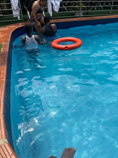 This image depicts a daytime scene at a swimming pool in Aba, Nigeria. Four individuals are present. One individual, wearing a dark bikini top and patterned shorts, is partially submerged near a metallic pool ladder at the pool's edge. Behind this individual, white towels are draped over a silver railing. Another individual, seen from behind and wearing a white tank top, is partially submerged, resting forearms on the pool's brown-tiled edge. A third individual, also seen from behind and wearing a black tank top, is submerged deeper within the pool. An orange ring buoy floats in the clear blue water. The feet of a fourth individual are visible in the foreground, submerged in the water. The pool features blue mosaic tiles on its interior walls and bottom. Green foliage and a metal fence are visible in the background beyond the pool's edge.