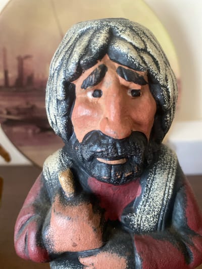 A close-up, head-on, eye-level shot features a small, vintage, painted figurine of a man. The figurine has a round face with a prominent nose, a black beard and mustache, and dark hair with gray streaks. The figure wears a red and black garment, with a gold-colored detail on its chest. The figurine is positioned in the foreground, with a blurred background that includes a circular, sepia-toned image of what appears to be industrial structures, possibly in a city setting. The lighting is bright and diffused, highlighting the textured surface of the figurine. The exact location in Almaty, Kazakhstan, is not discernible from the visual information provided.