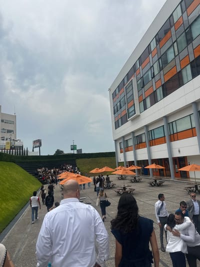 The image shows an outdoor scene at what appears to be a graduation ceremony, possibly in the city of San Bernardino Tlaxcalancingo, Mexico. A large group of people, many in white shirts, are seated in rows, likely graduates. Orange umbrellas provide shade. The background includes a modern building with large, rectangular windows and a white exterior. The sky is overcast. The foreground features a walkway where people in formal attire are congregated. Some individuals are embracing and interacting. A section of green lawn and a high fence run parallel to the seating area. A billboard is visible in the distance. The image is slightly angled.