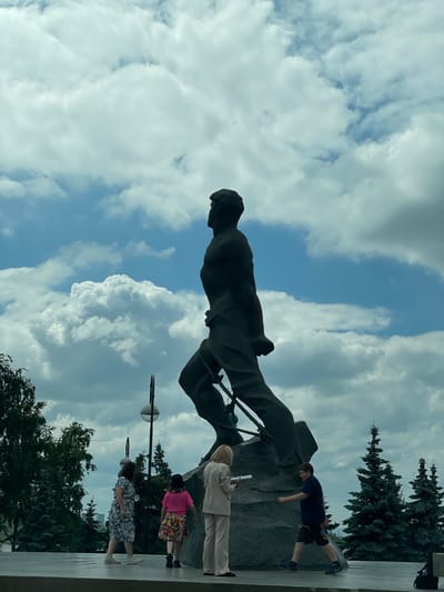 A low-angle shot captures a monumental bronze statue of a male figure in mid-stride against a dynamic cloudy sky in Kazan, Russia. The statue, a man with a determined expression and powerful build, is depicted with legs outstretched and arms bent, suggesting movement and strength. The statue is positioned on a large, dark-colored rock base, with details of its construction and rigging visible on its legs.

In the foreground, four individuals are observed. On the left, a woman in a blue and white floral dress walks away from the camera. To her right, a young person in a pink shirt and a colorful skirt walks towards the statue. Further to the right, a woman in a light-colored suit stands, looking down at some papers in her hand. On the far right, a young male wearing a dark blue polo shirt and patterned shorts walks towards the statue, seemingly engaged in a casual stroll.

The environment is characterized by lush green trees, including several tall fir trees, framing the right side of the image. A lamppost with a spherical light fixture is visible behind the woman in the floral dress. The ground surface is a light-colored, smooth pavement or platform. The sky is filled with dramatic cumulus clouds, creating a sense of grandeur and scale. The overall impression is one of an outdoor public space, likely a park or monument area, where people are enjoying the scenery and the presence of the imposing artwork.