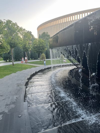 The image depicts a section of the Krasnodar Stadium (also known as the Kuban Stadium) park in Krasnodar, Russia. In the foreground, a modern, multi-tiered water feature with dark, sleek stone surfaces is visible. Water cascades down the steps of the fountain, creating a dynamic and reflective surface. Beyond the fountain, a pathway curves through a well-maintained park with lush green lawns and mature trees.

Several people are present in the park. Three women are walking on a paved path in the middle ground. Two of them are carrying bright orange bags. Further back, a young child stands on the pathway near the fountain, looking towards the water. In the background, a couple is seated on the grass, enjoying the park.

Dominating the upper portion of the image is the distinctive architectural structure of the Krasnodar Stadium. Its circular design and rhythmic vertical elements are clearly visible against a bright, overcast sky. The overall scene suggests a pleasant, leisurely day in a modern urban park setting, with the stadium serving as a prominent landmark.
