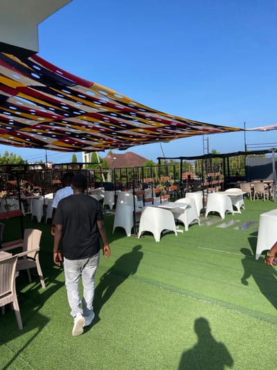 This image captures an outdoor leisure or event space under a clear, bright blue sky, likely during midday or early afternoon due to the strong sunlight and distinct shadows. The ground is covered with artificial green turf.
In the foreground, a person is seen from behind, wearing a black short-sleeved shirt, light grey pants, and white sneakers, standing on the turf. Another person's forearm and hand, holding a brown object, are visible on the right. Long, pronounced shadows of individuals and objects stretch across the green surface.
The midground features rows of modern, white, possibly plastic or rattan-style chairs, neatly arranged on the turf. Beyond these, additional darker, wicker-style chairs and tables are visible. A black metal railing or fence runs horizontally across the midground, with small potted plants placed on a ledge behind it.
A prominent feature in the left and upper parts of the frame is a large, colorful fabric sunshade or awning. It displays a vibrant geometric pattern of red, yellow, black, and light-colored stripes and shapes, interspersed with numerous circular cutouts that allow light to filter through. The background shows glimpses of a brown-tiled roof and some distant greenery, hinting at surrounding structures or natural elements.
The setting appears to be a rooftop lounge or an elevated outdoor venue, designed for comfort and social gatherings, consistent with a sunny location such as Dar es Salaam, Tanzania. No visible text is present in the image.
