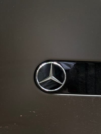 The image displays a close-up view of a Mercedes-Benz logo emblem on the exterior of a vehicle. The emblem, a silver three-pointed star within a silver circle, is positioned on a black horizontal trim piece. The vehicle's body is a dark, matte color, possibly a deep brown or grey, and appears to have a textured surface with some minor imperfections. The lighting suggests an outdoor setting, with highlights reflecting on the chrome of the emblem and the glossy black trim. The overall context of the image is a still shot of a car's branding, with no indication of a specific urban location or environment. The image does not provide any information that can be used to identify its location in Belgrade, Serbia.